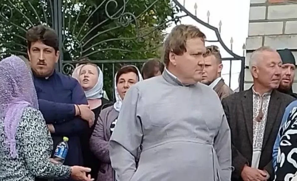 Група людей і священник у сірій рясі біля кованої брами храму
